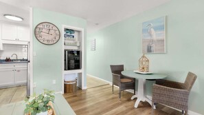 A room with a dining area, a clock, a refrigerator, and a wicker chair. - Shore Thing - Studio Stay (Panama City Beach)