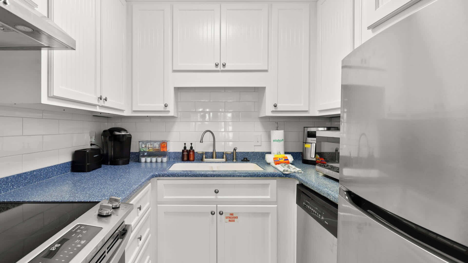 Uma cozinha moderna com armários brancos, bancada azul, cafeteira, pia e máquina de lavar louça.