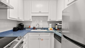 A modern kitchen with white cabinets, a blue countertop, a coffee maker, a sink, and a dishwasher. - Shore Thing - Studio Stay (Panama City Beach)
