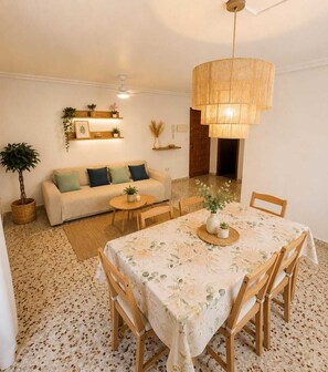 Ein gemütliches Wohnzimmer mit einer Couch, einem runden Tisch mit Stühlen, einer Hängelampe und einer Tür im Hintergrund.