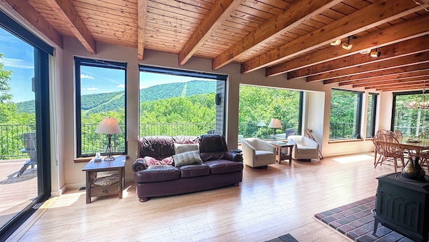 A spacious living area with a wooden ceiling, large windows offering a view of green hills, a leather sofa, and a dining table with chairs.
