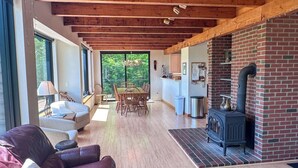 A spacious living area with a wooden ceiling, a dining table with chairs, a fireplace, and a view through large windows.