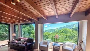 A spacious living room with a wooden ceiling, large windows offering a view of a forested hillside, and a giraffe figurine near the window.