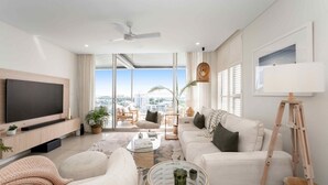 A modern living room with a flat-screen TV, comfortable seating, a glass coffee table, and a city view through large windows. - Absolute Waterfront Luxe Sub-Penthouse | 3BR | Panoramic Views (South Townsville)