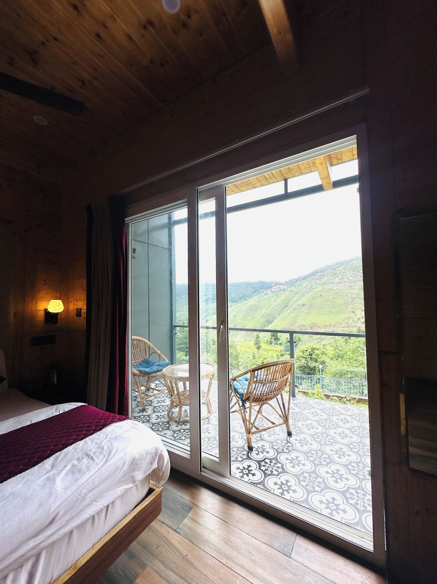 A bedroom with a bed, a nightstand, a lamp, and a view of a balcony with chairs and a scenic landscape.