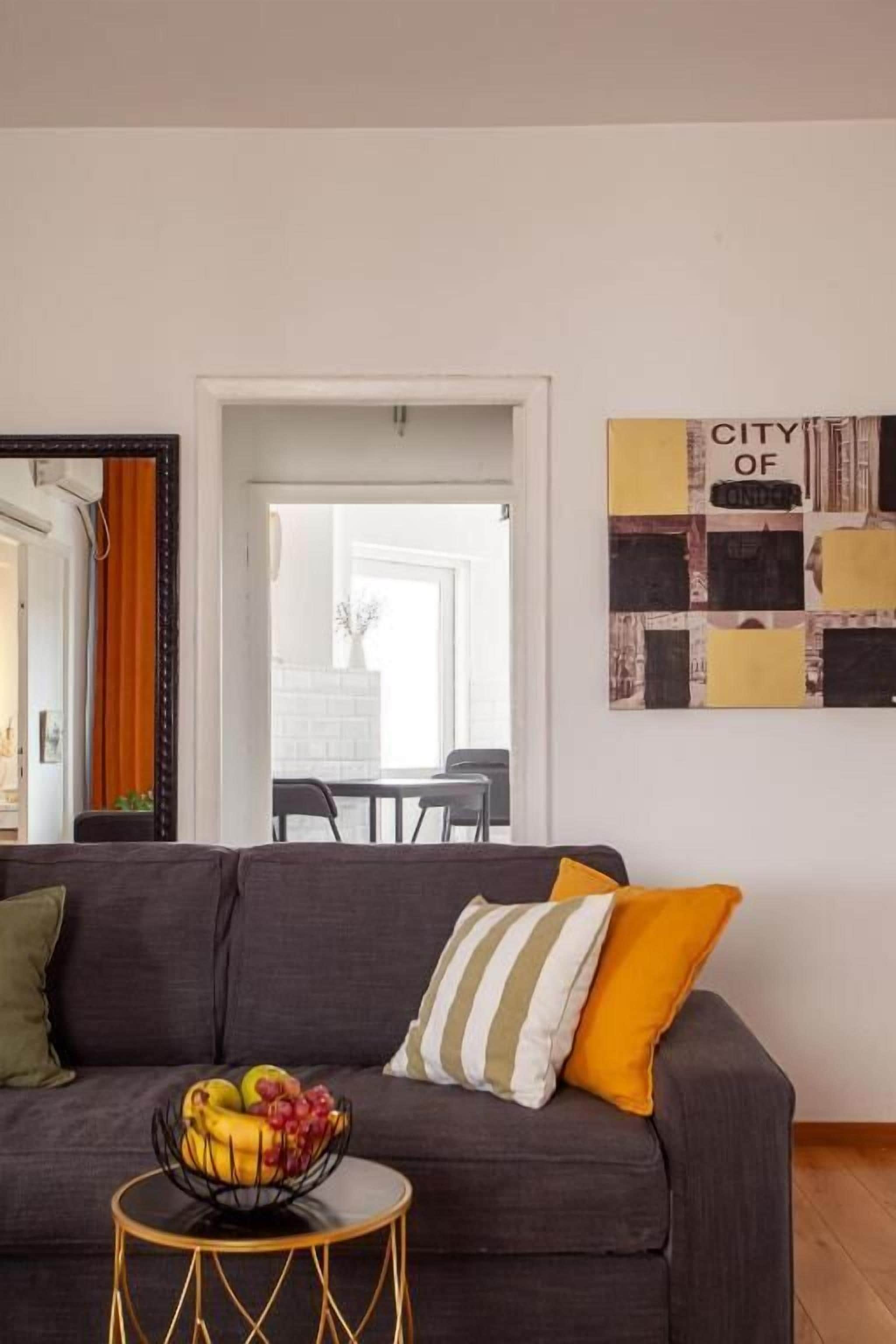 Un salón con un sofá, una mesita redonda con un frutero, y una pared decorada con un collage de arte.