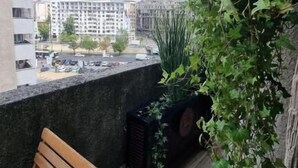 Un balcón con una silla de madera, una maceta negra y vistas a edificios bajo un cielo despejado.