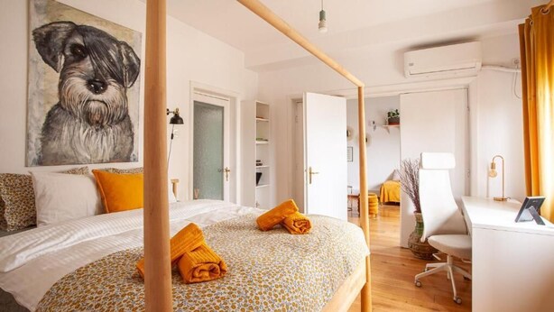 Un dormitorio con cama, escritorio y silla. Hay un cuadro en la pared y una puerta que da a otra habitación.