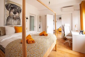 Quarto com cama, escrivaninha e cadeira. Há um quadro na parede e uma porta que dá para outro cômodo.