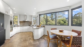 A modern kitchen with white cabinetry, a central island, and a dining area with a round table and woven chairs. - Riverfront Alpine Retreat - Mt Buller Gateway - Sleeps 10 (Merrijig)