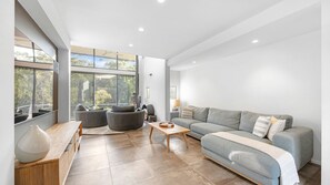 A spacious living area with a grey sofa, a wooden coffee table, and a large window offering a view of trees. - Riverfront Alpine Retreat - Mt Buller Gateway - Sleeps 10 (Merrijig)
