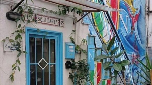 Un edificio con puerta azul, balcón con barandilla metálica y un mural colorido en la pared.