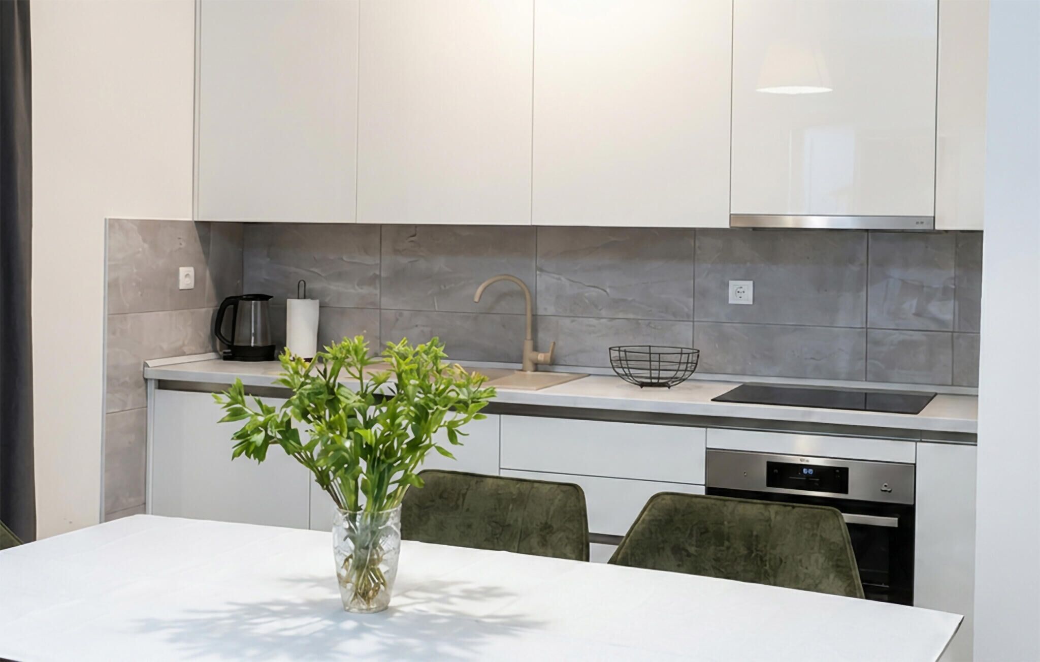 A modern kitchen with a white countertop, a sink, a kettle, and a vase with green plants.