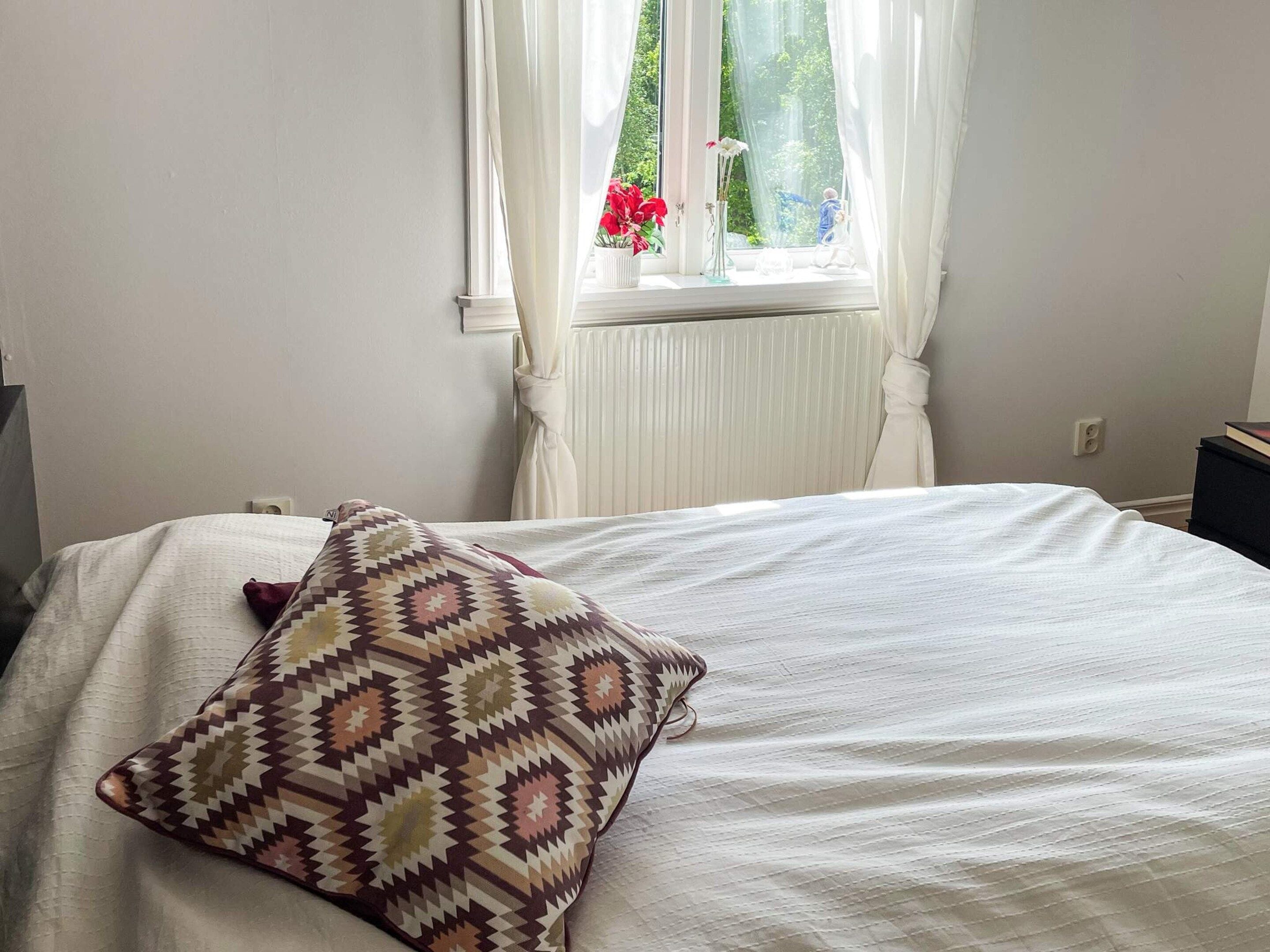Un dormitorio con cama, ventana con cortinas, un radiador y una pequeña mesa con un libro.
