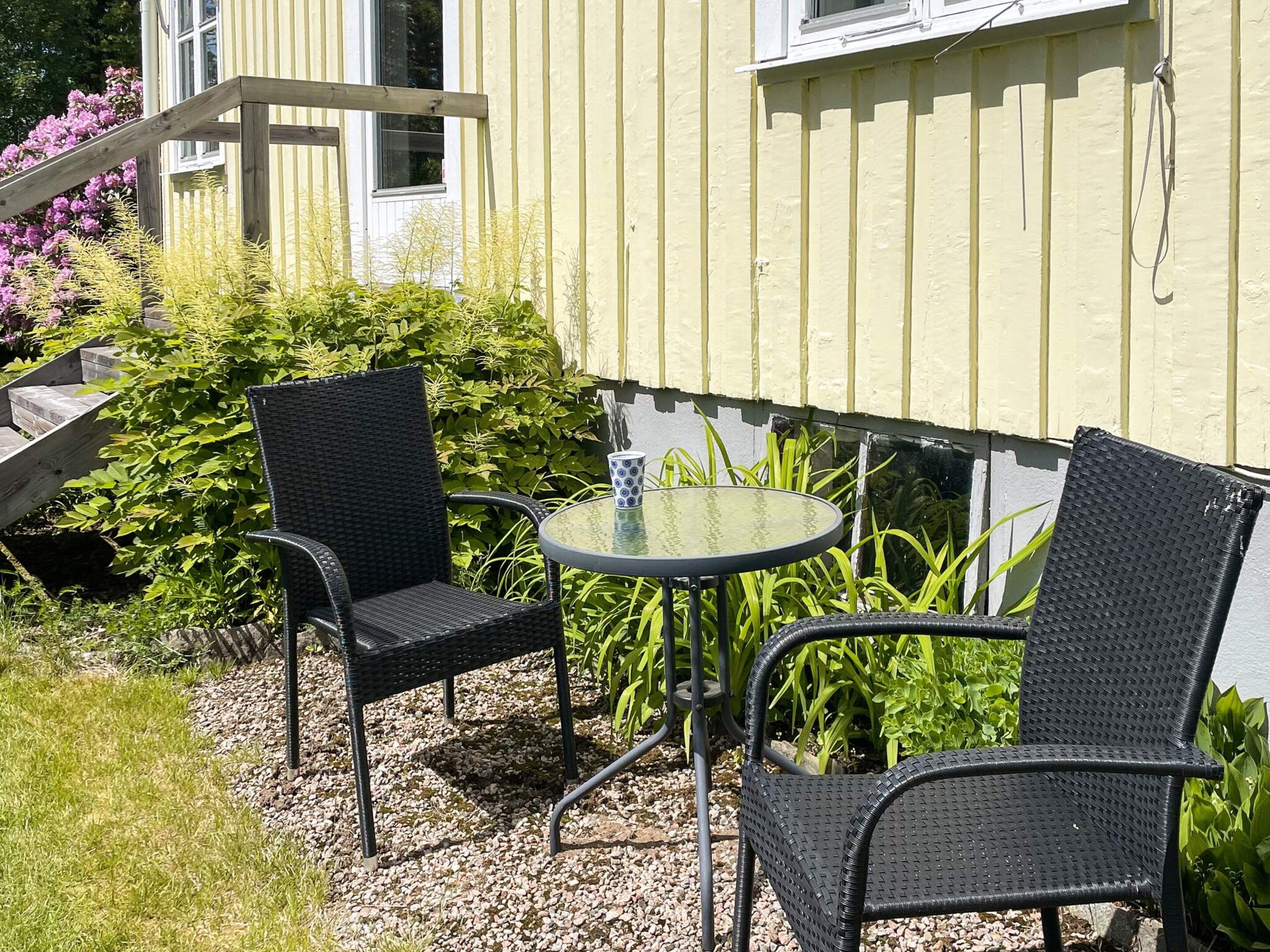 An outdoor seating area with two chairs and a table, surrounded by greenery and a building with a yellow exterior.