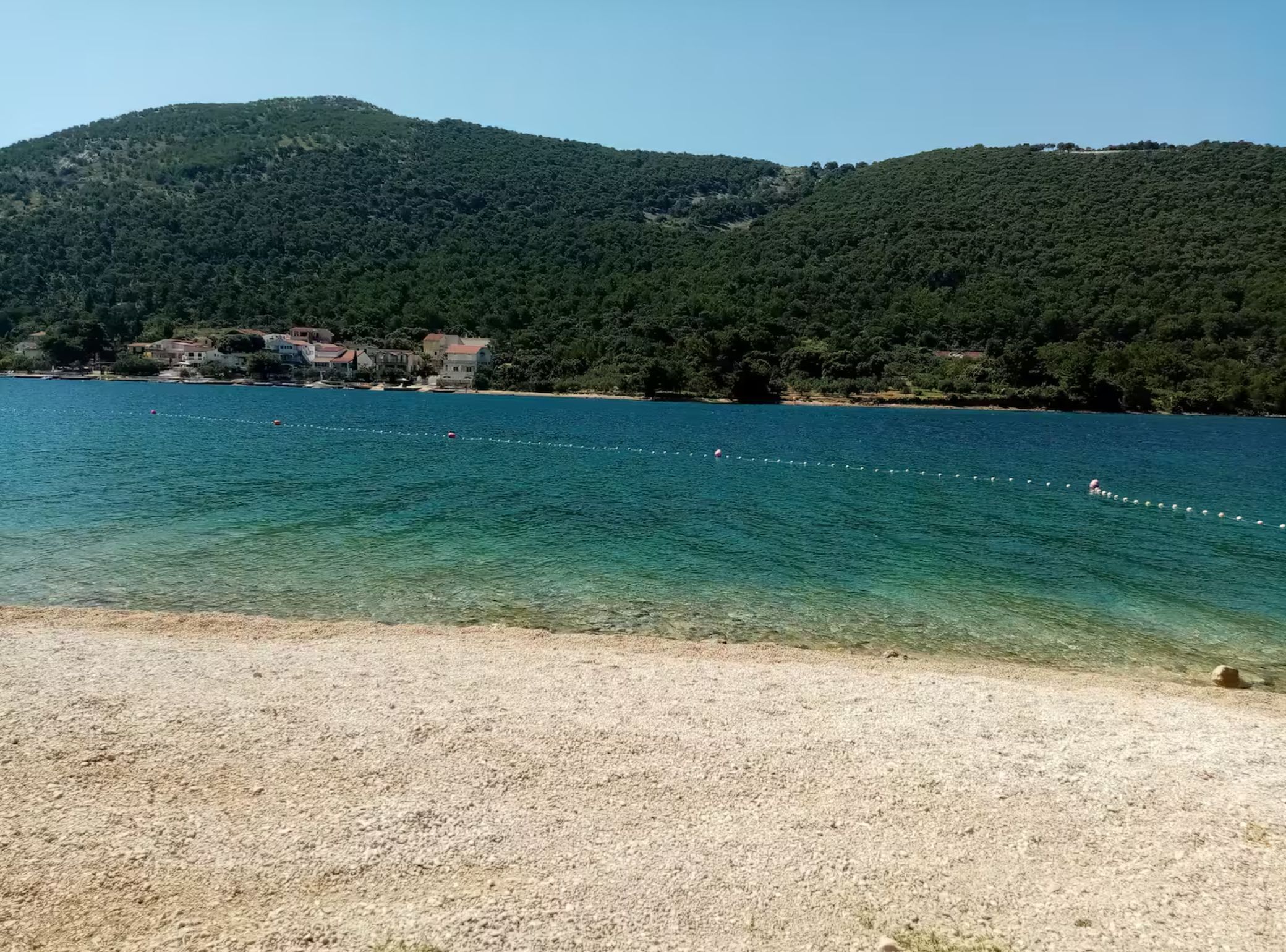 A beach with clear water, a sandy shore, and a backdrop of hills with buildings.