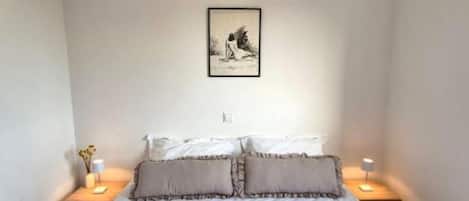 A bedroom with a bed, two bedside tables with lamps, a framed picture on the wall, and a small vase on one table.