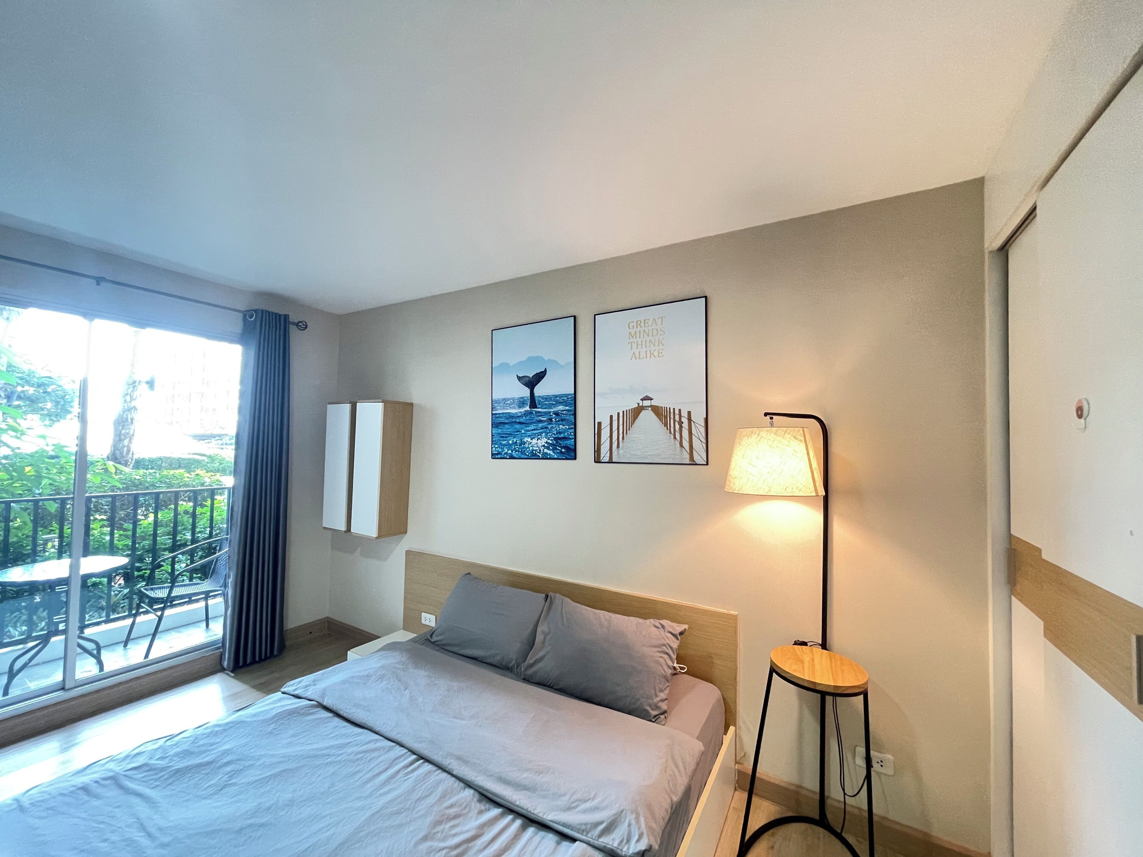 A hotel room with a bed, a bedside lamp, a small table, a window with a view of greenery, and two wall-mounted pictures.