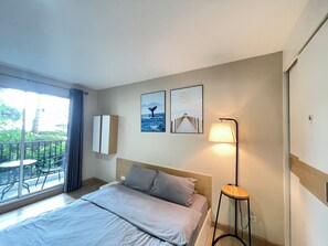 A hotel room with a bed, a bedside lamp, a small table, a window with a view of greenery, and two wall-mounted pictures. - Room16atchangwattana (Pak Kret)