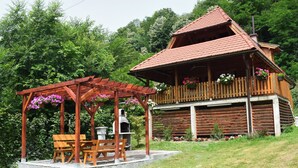 A wooden cabin with a balcony, a wooden pergola with flowers, and a barbecue grill.