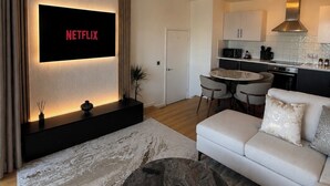 Uma sala de estar moderna com uma televisão de tela plana, um sofá, uma mesa de centro e uma cozinha ao fundo.