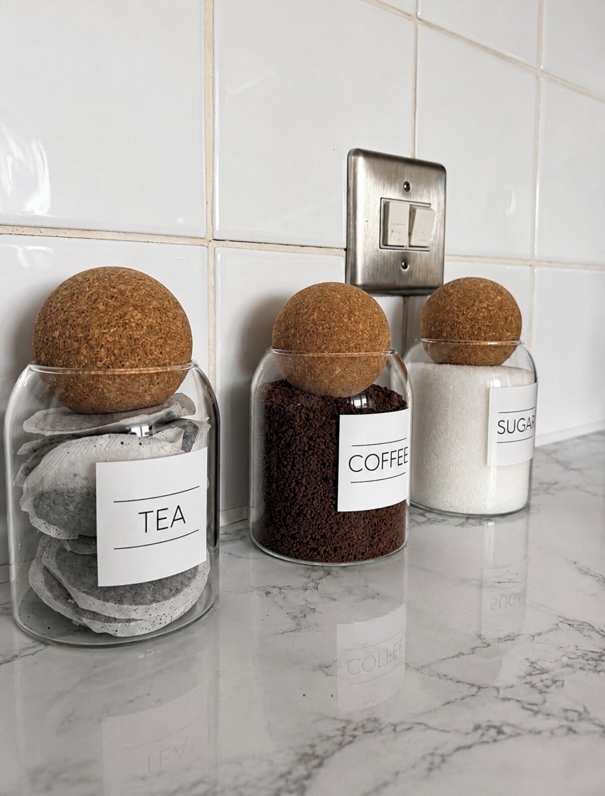 Trois bocaux en verre avec des couvercles en liège, posés sur un comptoir en marbre, et portant les étiquettes « THÉ », « CAFÉ » et « SUCRE ».