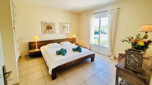 Ein Hotelzimmer mit einem Bett, zwei Nachttischlampen, einem Fenster mit Vorhängen, einem kleinen Tisch mit einer Lampe und zwei gerahmten Bildern an der Wand.