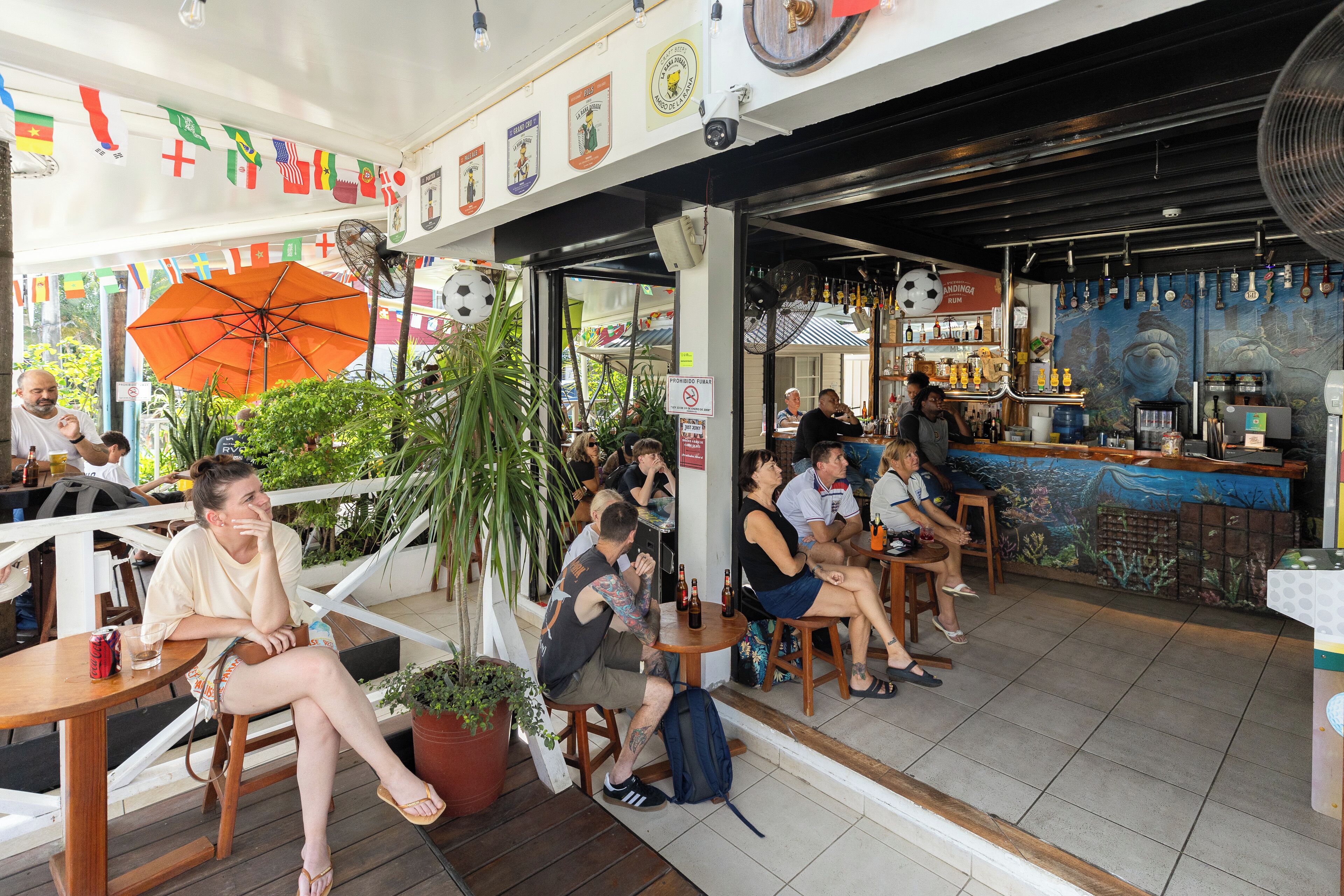 An outdoor seating area with tables and chairs, surrounded by potted plants and flags.