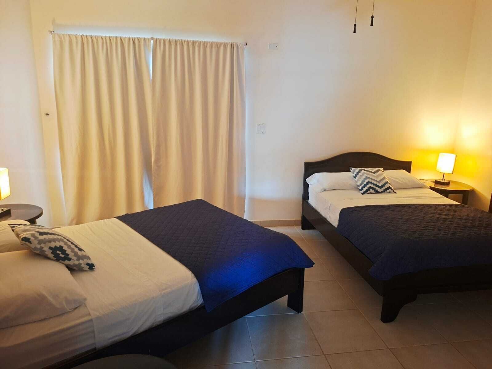 A hotel room with two beds, a window with curtains, a bedside lamp, and a tiled floor.