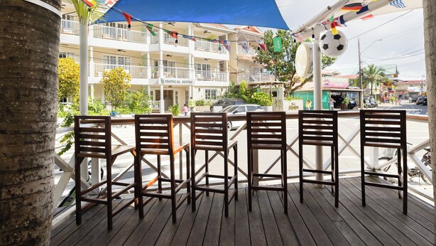 Terraza de madera con sillas, vista de un edificio de varios pisos con balcones y una calle con vehículos y peatones.