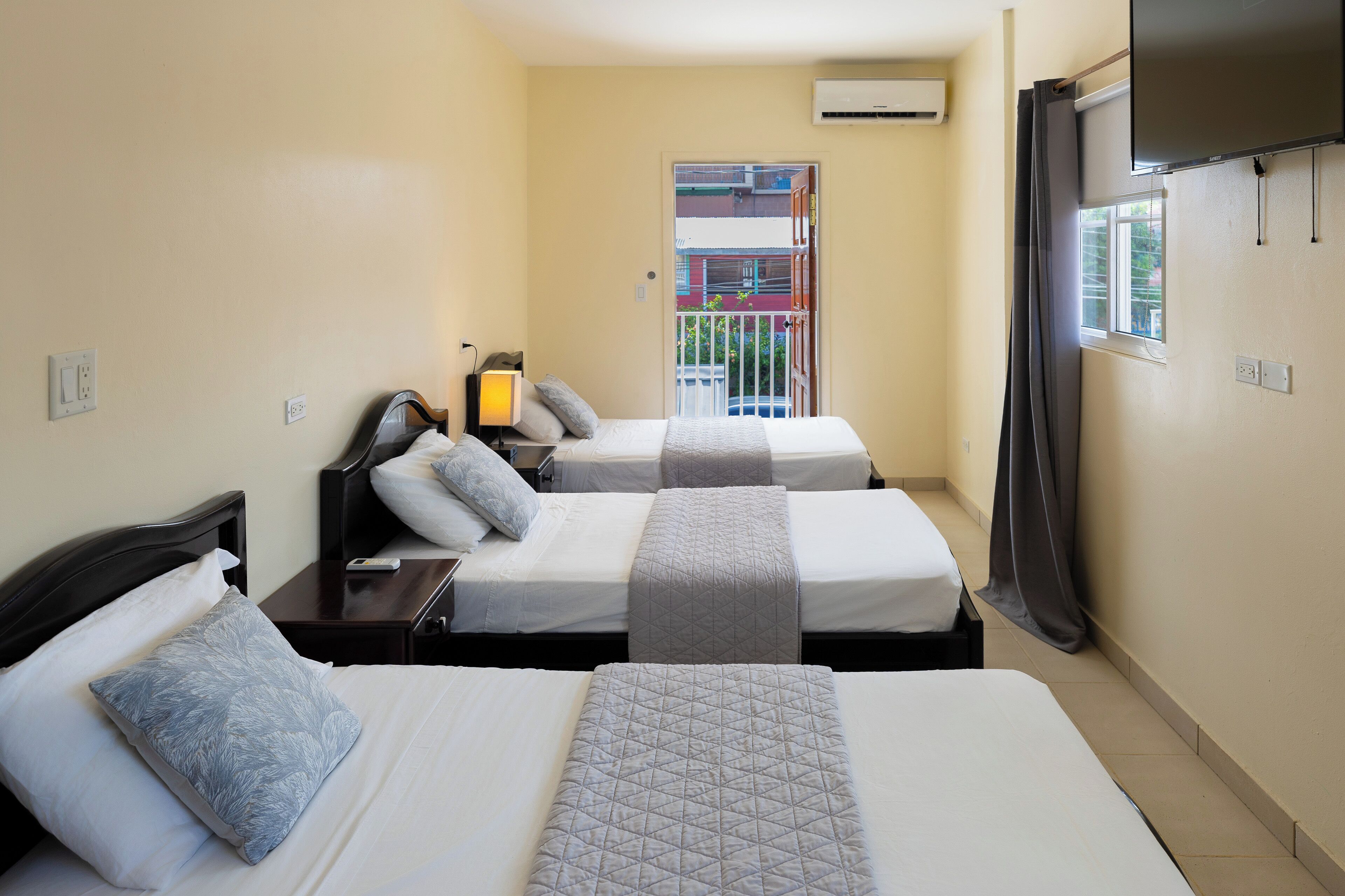 A hotel room with two beds, a bedside table with a lamp, a window with curtains, and an air conditioning unit on the wall.