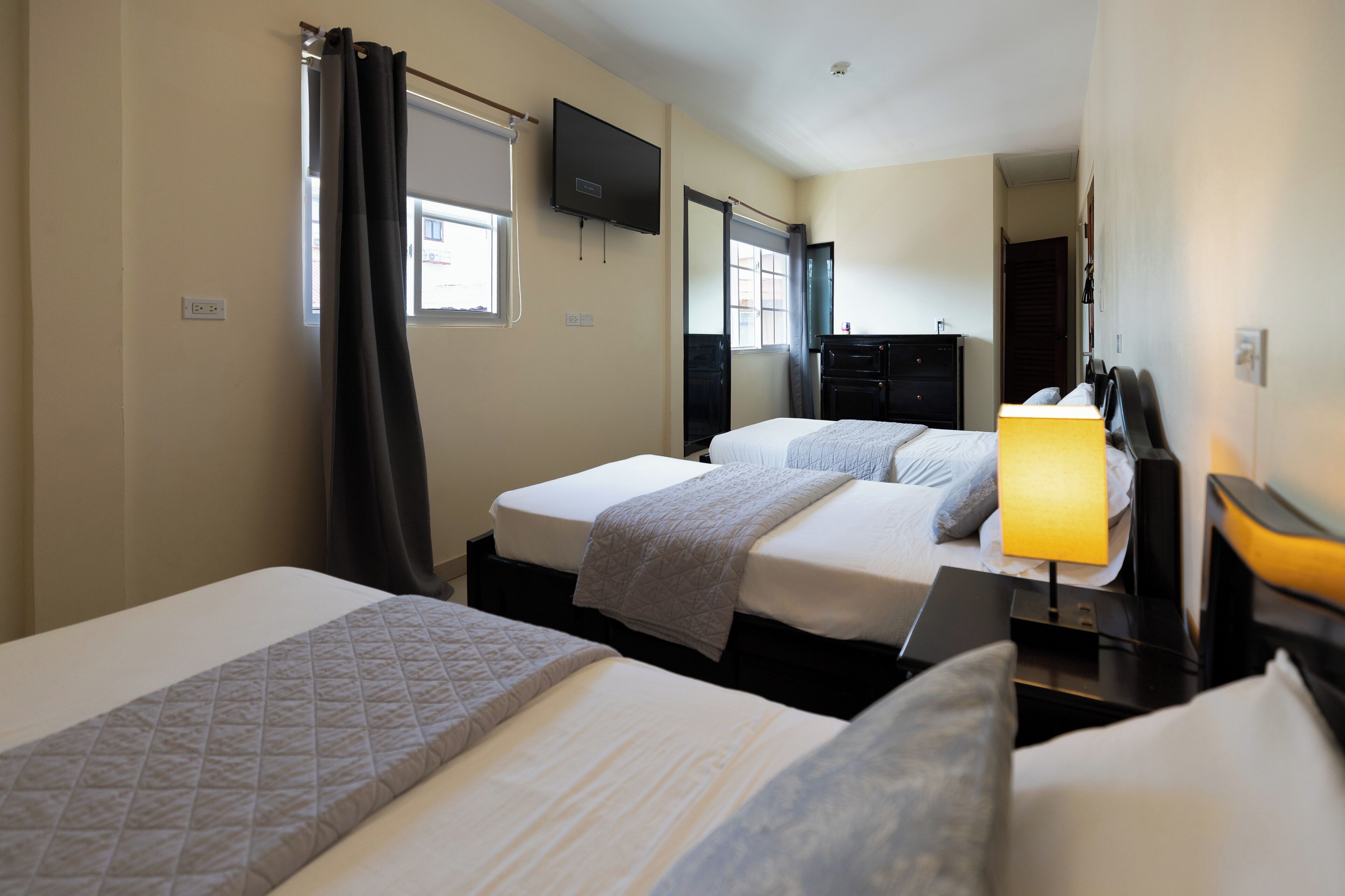 A hotel room with two beds, a flat-screen TV mounted on the wall, a desk with a lamp, and a window with curtains.