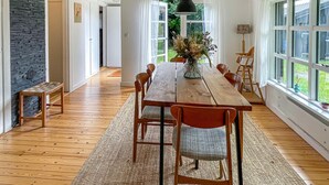 A dining area with a wooden table, chairs, a vase with flowers, and a view through large windows. - 2 bedroom stunning home in Rørvig (Rørvig)