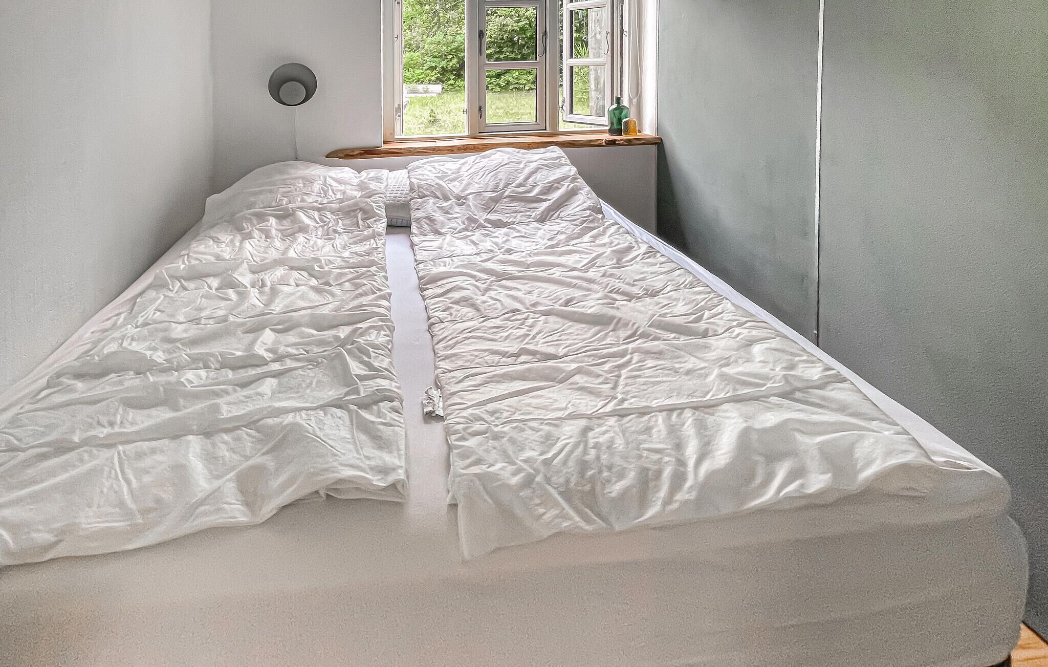 A single bed with a white mattress and a zipper, a window with a view of greenery, and a wall-mounted light fixture.