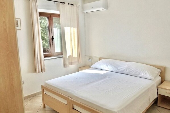 Ein Schlafzimmer mit einem Bett, einem Fenster mit Vorhängen, einem Nachttisch und einer Klimaanlage.