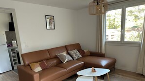 Een bruine leren bank met meerdere kussens, een witte salontafel en een tapijt met patroon in een kamer met een raam en een deur naar een keuken.