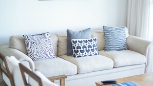 Una sala de estar con un sofá color beige, cojines estampados, sillas de madera, una mesa de centro y un cuadro con una playa.