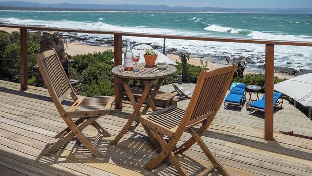 Terraza de madera con dos sillas y una mesa, con vista a una playa y olas.