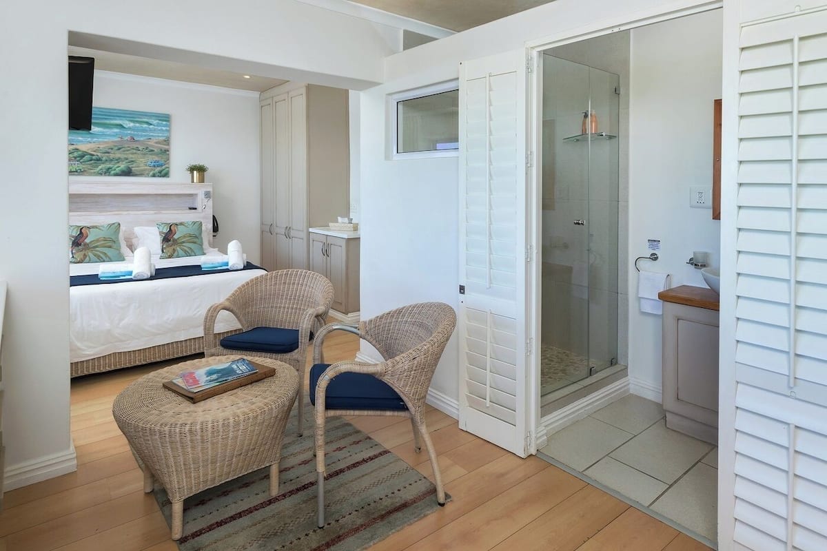 A bedroom with a bed, two wicker chairs, a small table, a painting, and a glass-enclosed shower.