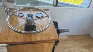 Una rueda de timón de madera en la cubierta de mando de un barco, una ventana con vistas al mar, y un panel de control con varios diales e interruptores.