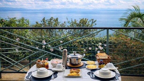 Una mesa lista para el desayuno con vistas al mar, una baranda de metal y follaje al fondo.