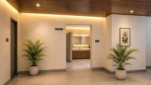 A hallway with a sign indicating a washroom, potted plants, and a glimpse of a bathroom with a sink and mirror. - Yashshree Namdang Premier Hotel & Banquet (Margherita)