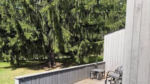Eine Holzterrasse mit Metallstühlen, eine Betonwand und ein großer Baum im Hintergrund.