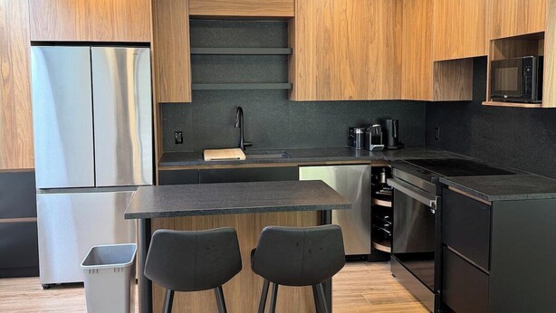 Eine moderne Küche mit Holzschränken, einem Edelstahlkühlschrank, einer schwarzen Arbeitsplatte und zwei schwarzen Barhocker.