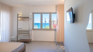 Quarto de hotel com cama, escrivaninha, televisão fixada na parede e janela vista para o mar.