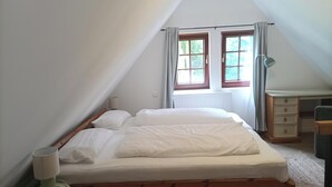 Ett sovrum med ett sluttande tak, två fönster med gardiner, en säng med vita sängkläder, en sänglampa och en byrå med lådor.