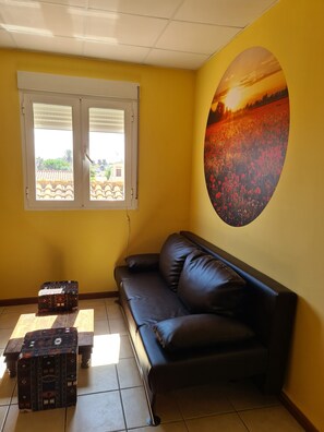 A room with a yellow wall, a black leather sofa, a small patterned ottoman, a window with a view of buildings, and a large oval painting of a sunset. - A Little Getaway - El Refugio Amarillo, Studio for Three, Near the Beach (Els Poblets)