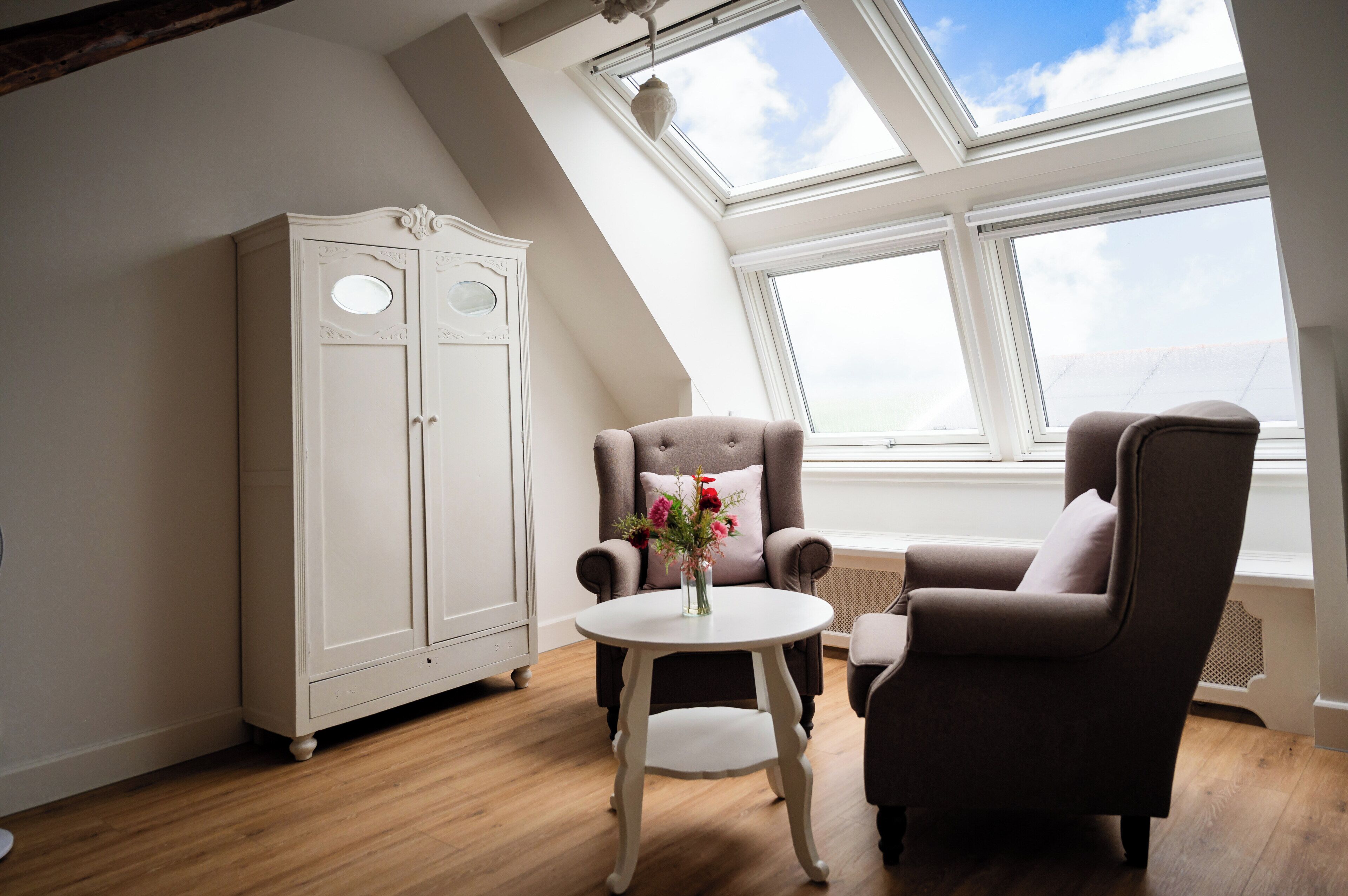 A room with a sloped ceiling, a white wardrobe, two armchairs, a small round table with a vase, and large windows.