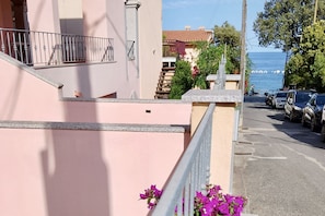 A pink building with a balcony, a street with parked cars, and a view of the sea. - Cala Gonone, 2-Bedroom Apartment (Cala Gonone)