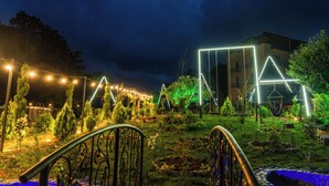 Un jardín con cerca de metal, terraza de madera y un edificio con formas geométricas iluminadas.